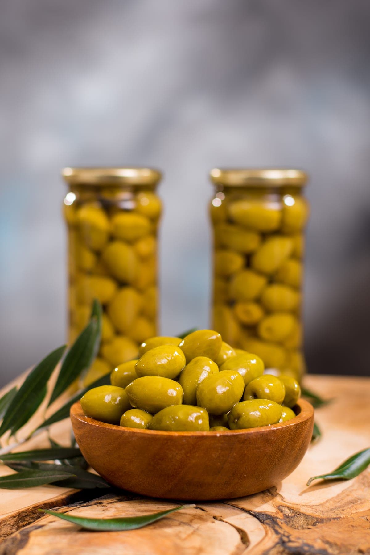 Frascos de aceitunas verdes junto a un bowl de madera con rama de olivo